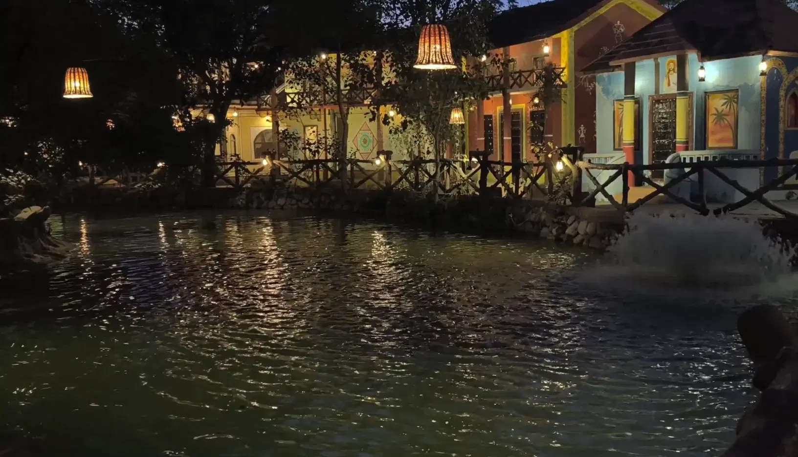Chokhi Dhani's illuminated traditional Rajasthani village resort at night with a decorative pond, hanging lanterns, and warm-lit heritage buildings reflected in the water, Jaipur